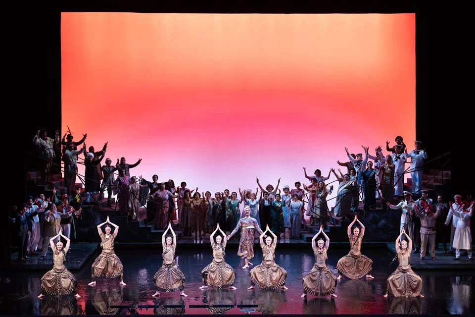Dancers in gold costumes kneeling in a row before the chorus, under a vast orange-pink backdrop – Lakmé at the Royal Opera House Muscat, Oman, 2019 – set design