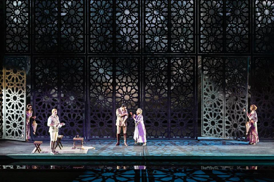 Singers before tall black screens with geometric lattice patterns, reflected in a wet stage floor – Lakmé at the Royal Opera House Muscat, Oman, 2019 – set design