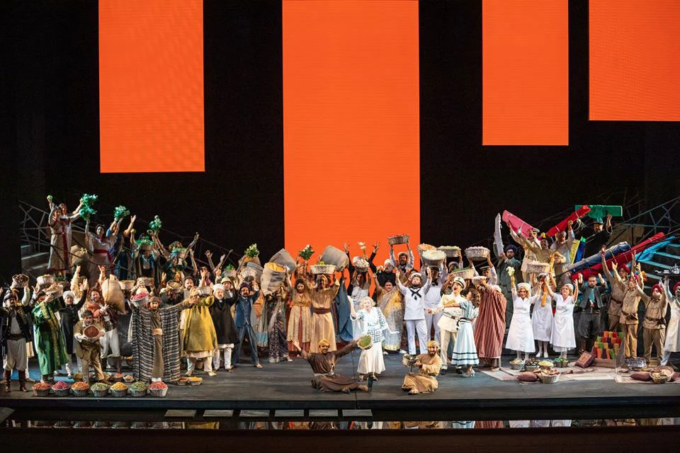 Crowded market scene with chorus holding baskets and goods under large orange panels – Lakmé at the Royal Opera House Muscat, Oman, 2019 – set design