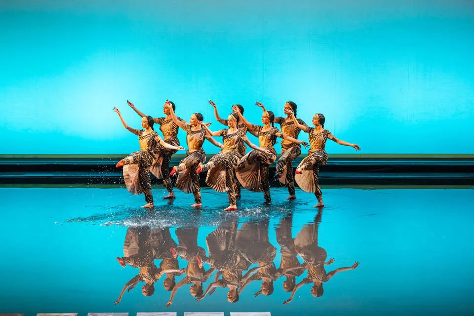 Dancers in patterned costumes splashing on a water-covered stage, reflected against a turquoise backdrop – Lakmé at the Royal Opera House Muscat, Oman, 2019 – set design