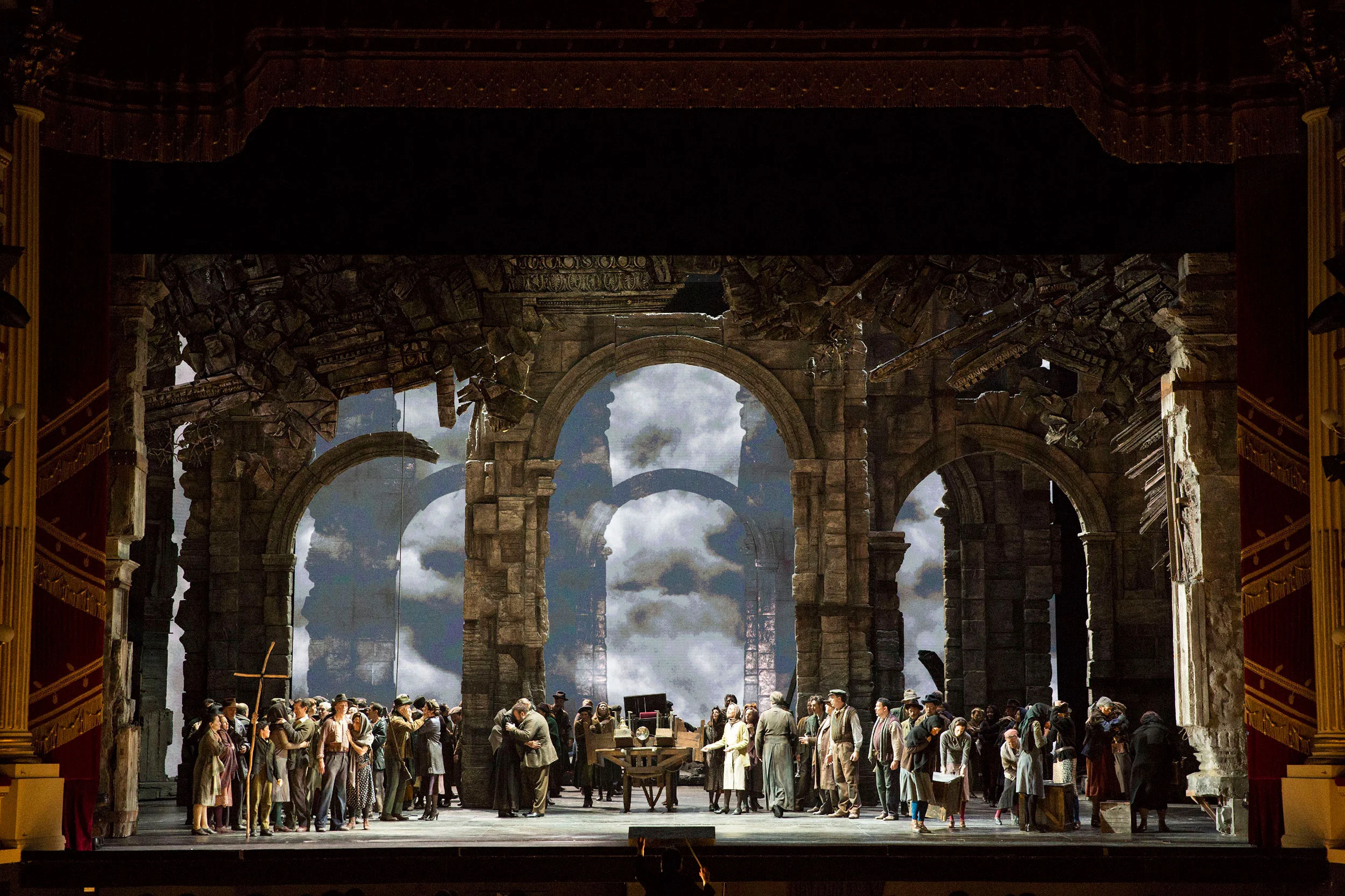 Crowd of villagers in a ruined Roman setting with stone arches open onto a cloudy sky – Attila at La Scala, Milan, Italy, 2018 – opera set design