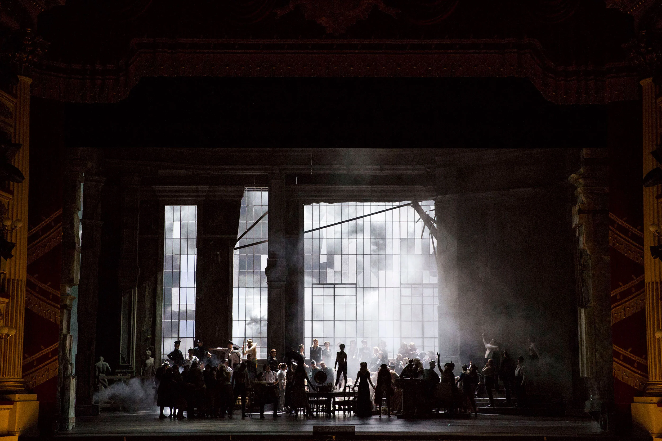 Crowd in silhouette before a bright shattered window, smoke drifting through the dark hall – Attila at La Scala, Milan, Italy, 2018 – opera set design