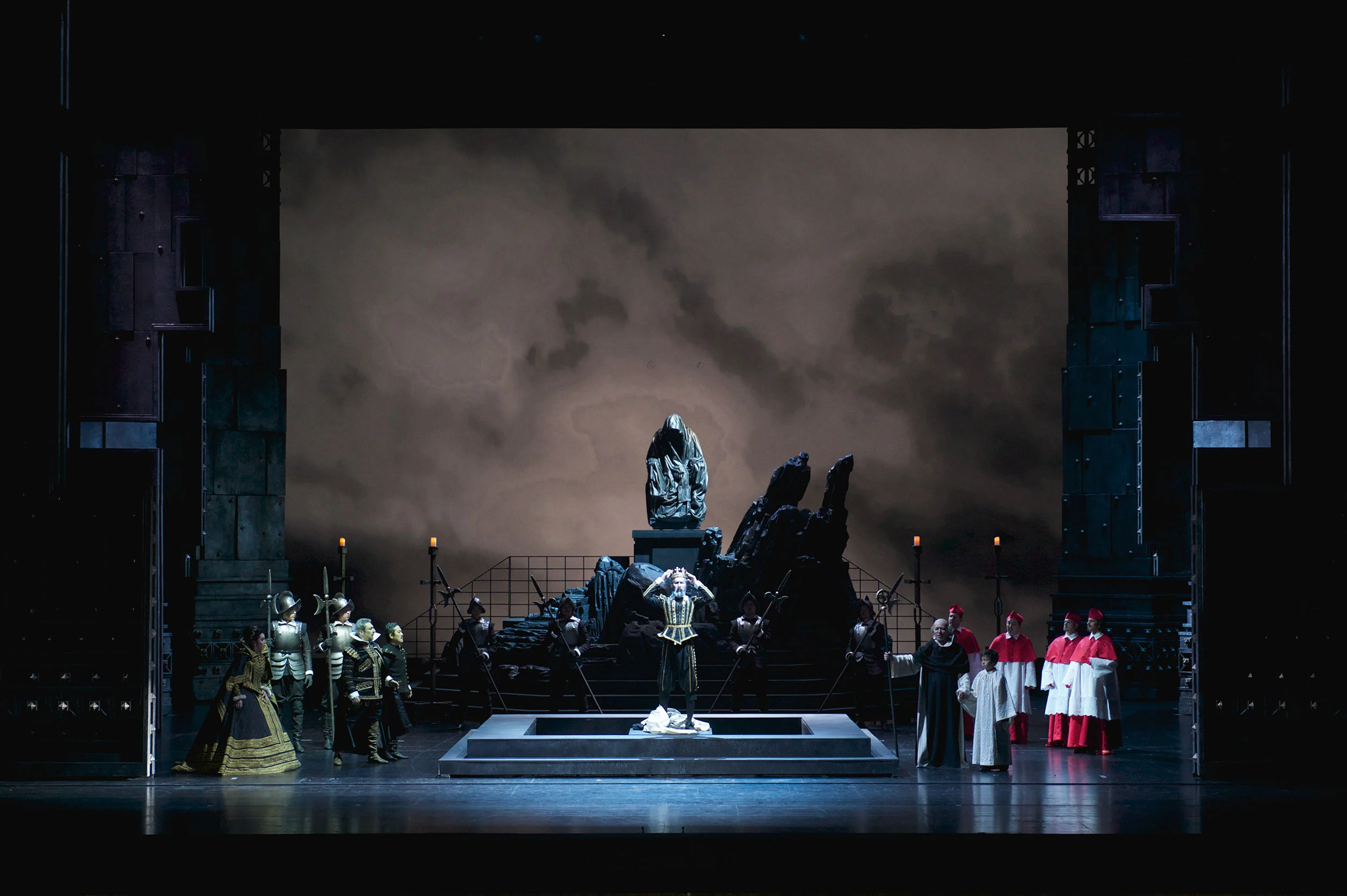 Singer kneeling on a dais before a stone statue, cardinals and soldiers either side under a cloudy sky – Don Carlo at Astana Opera, Kazakhstan, 2023 – set design