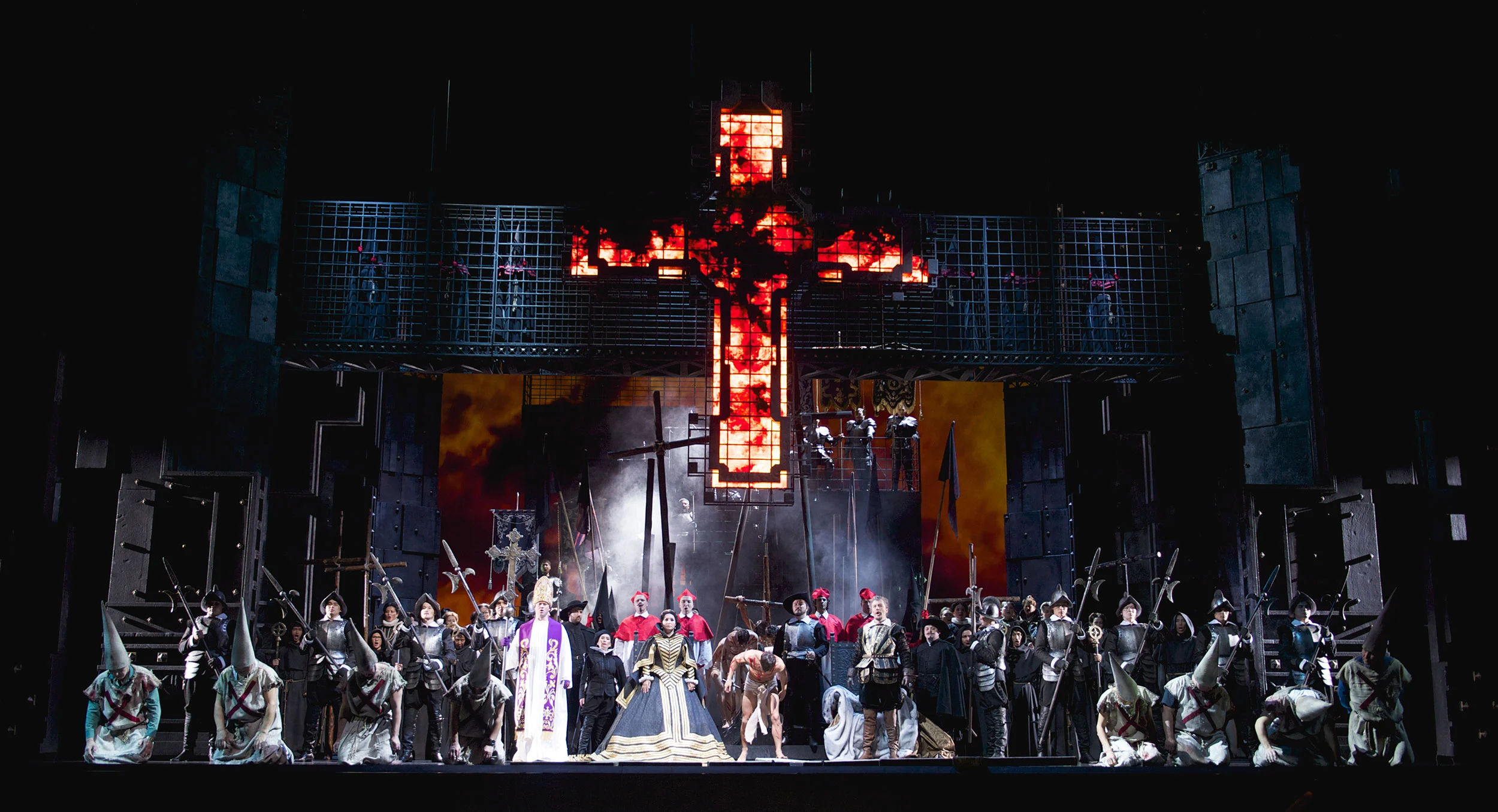 Full-cast finale before a burning cross, with hooded penitents in front of a grid of iron cages – Don Carlo at Astana Opera, Kazakhstan, 2023 – set design