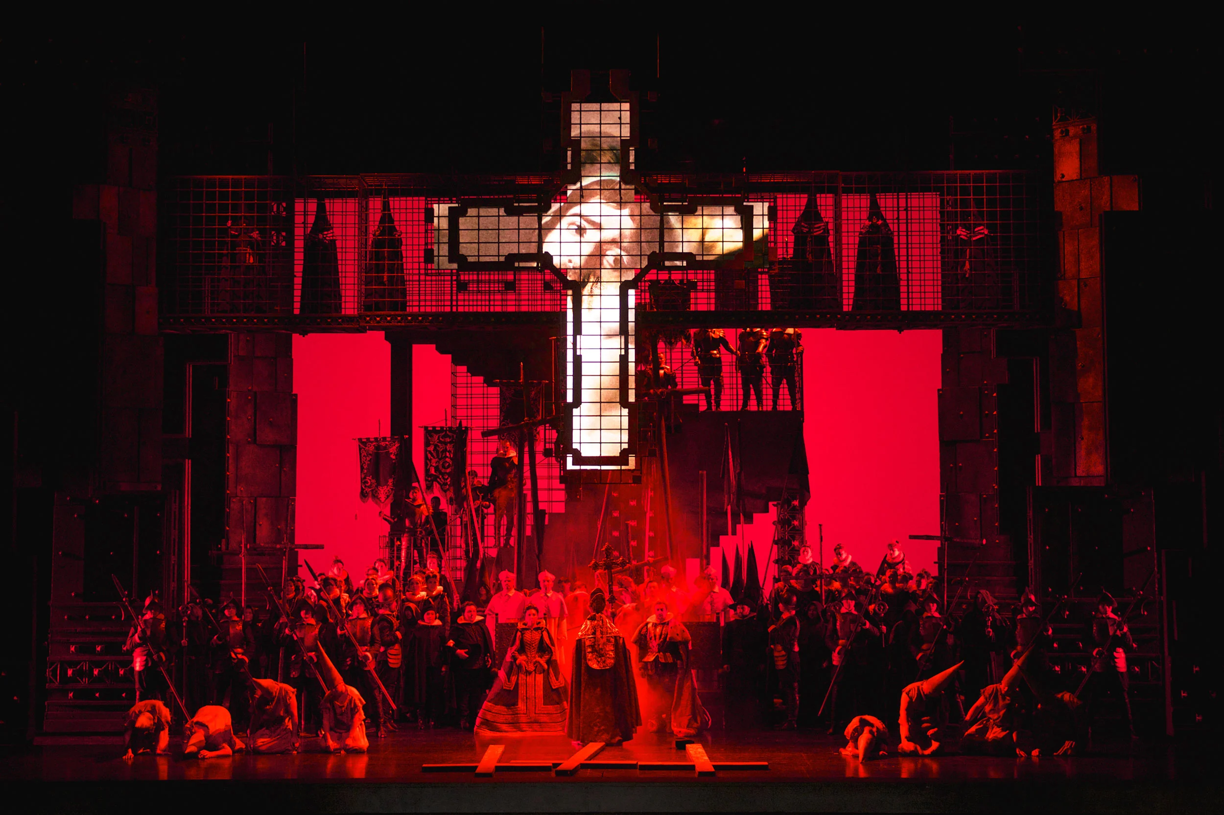 Stage flooded with red light below a giant cross of light, the chorus massed before the cage structure – Don Carlo at Astana Opera, Kazakhstan, 2023 – set design