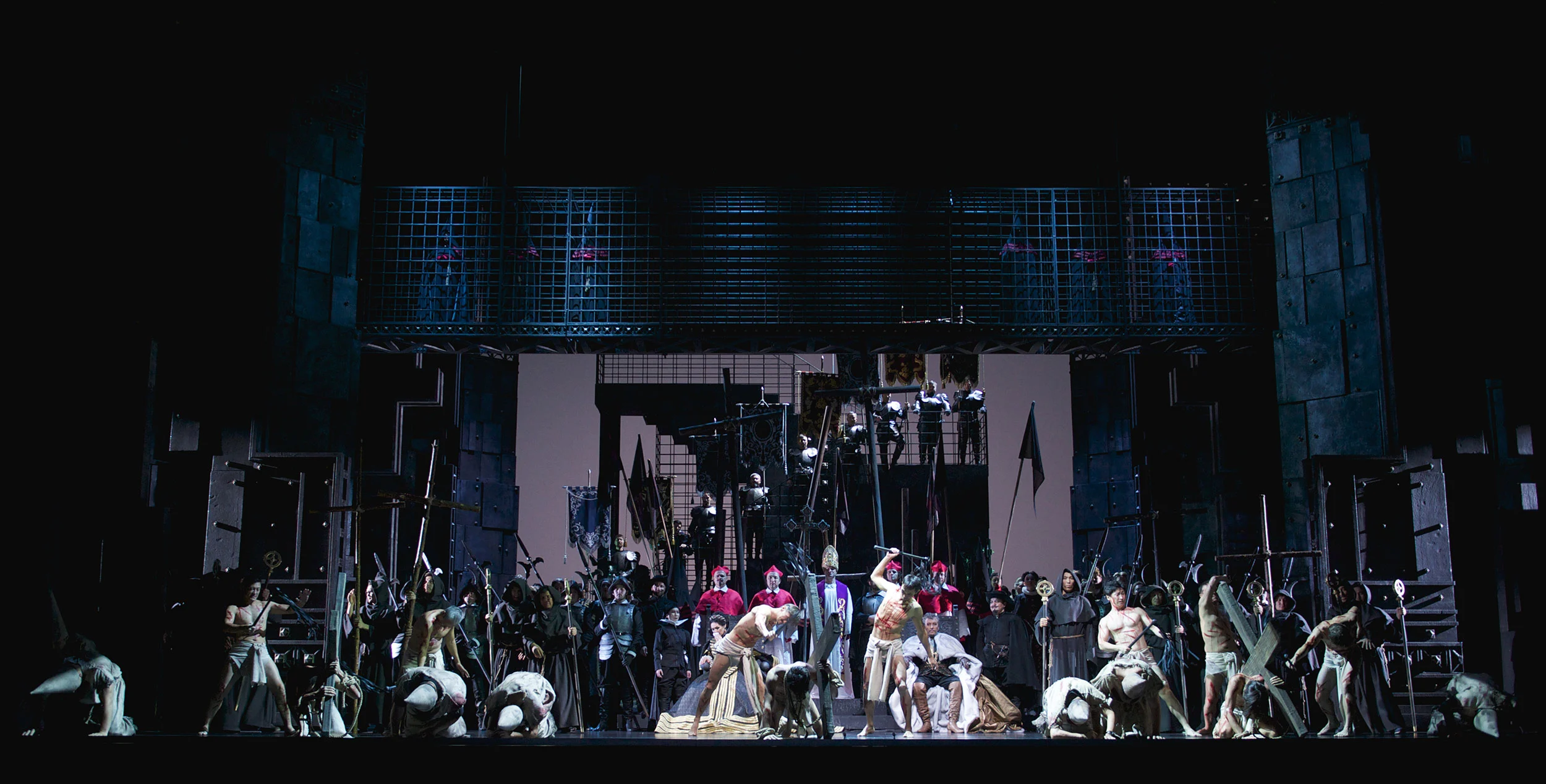 Wide view of a crowd scene with flags and soldiers under the iron cages, figures kneeling in front – Don Carlo at Astana Opera, Kazakhstan, 2023 – set design