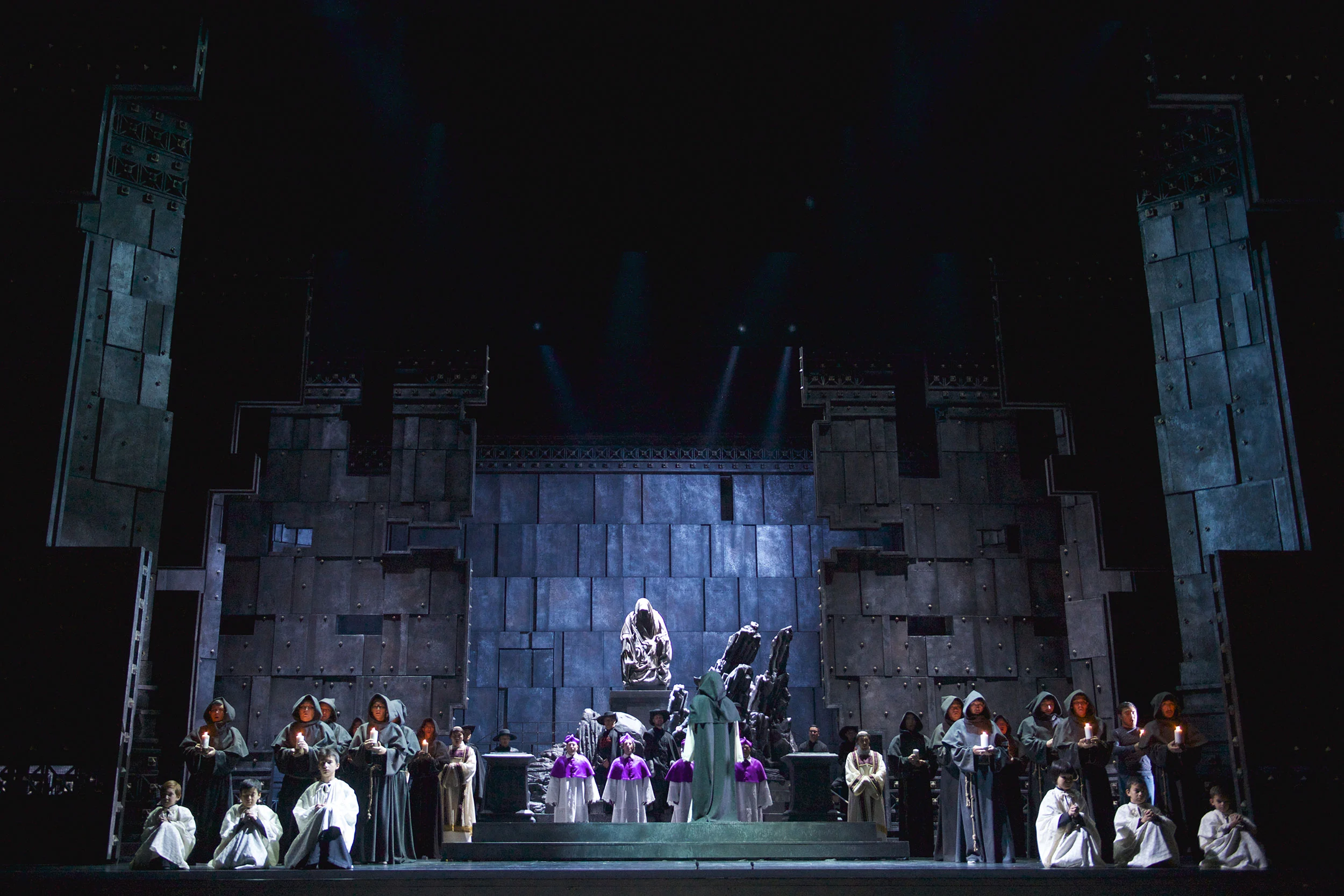 Monks with candles and a statue at centre before a monumental grey stone wall set – Don Carlo at Astana Opera, Kazakhstan, 2023 – set design