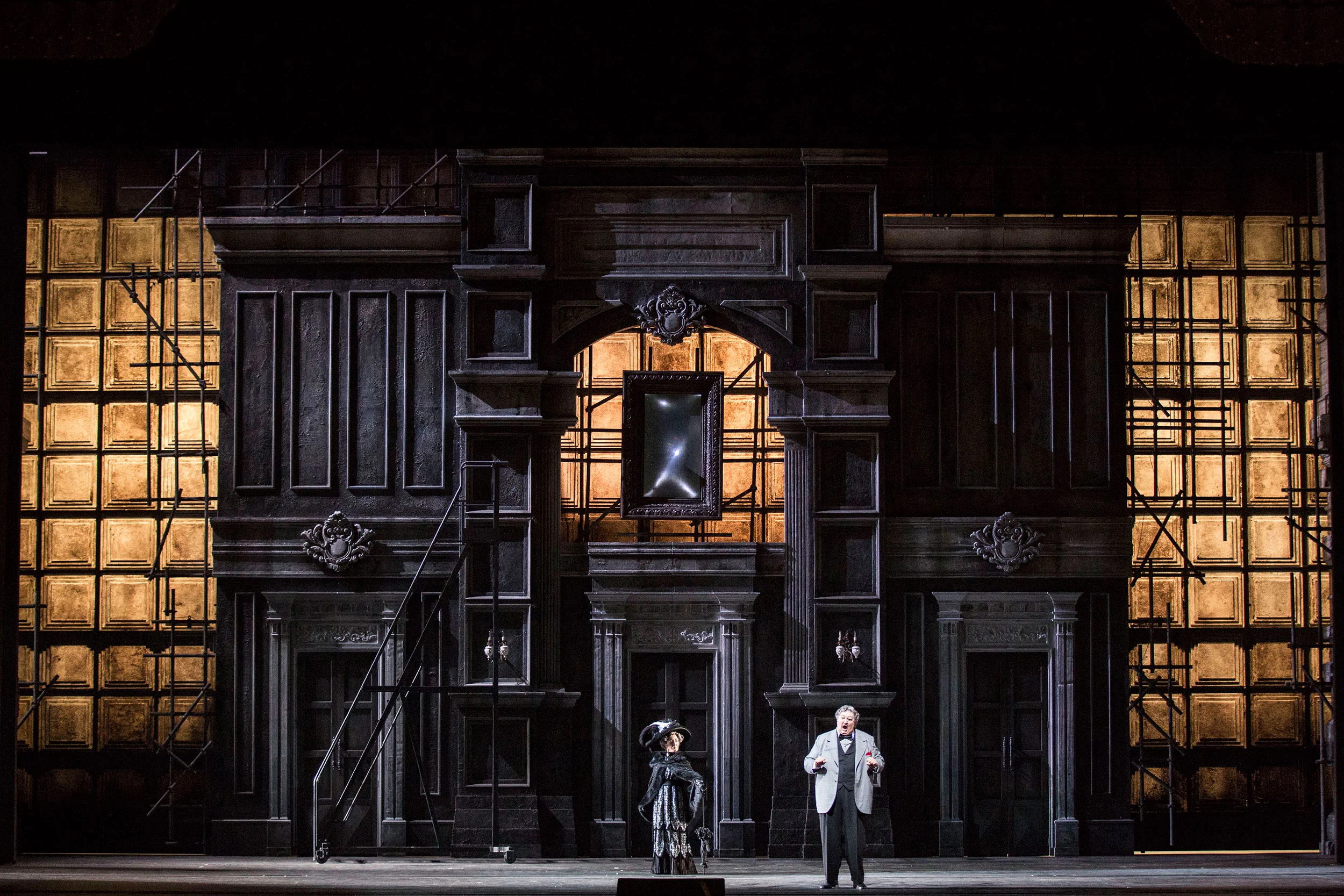 Two singers before a grey carved facade framed by scaffolding with warm lit windows – Don Pasquale at La Scala, Milan, Italy, 2018 – opera set design