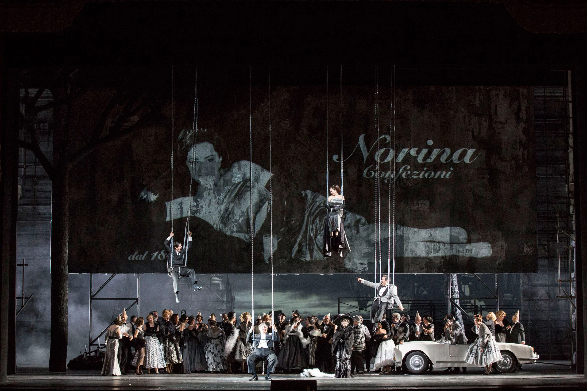 Chorus on stage with swings hanging before a projection of a woman and the words Norina Confezioni – Don Pasquale at La Scala, Milan, Italy, 2018 – opera set design