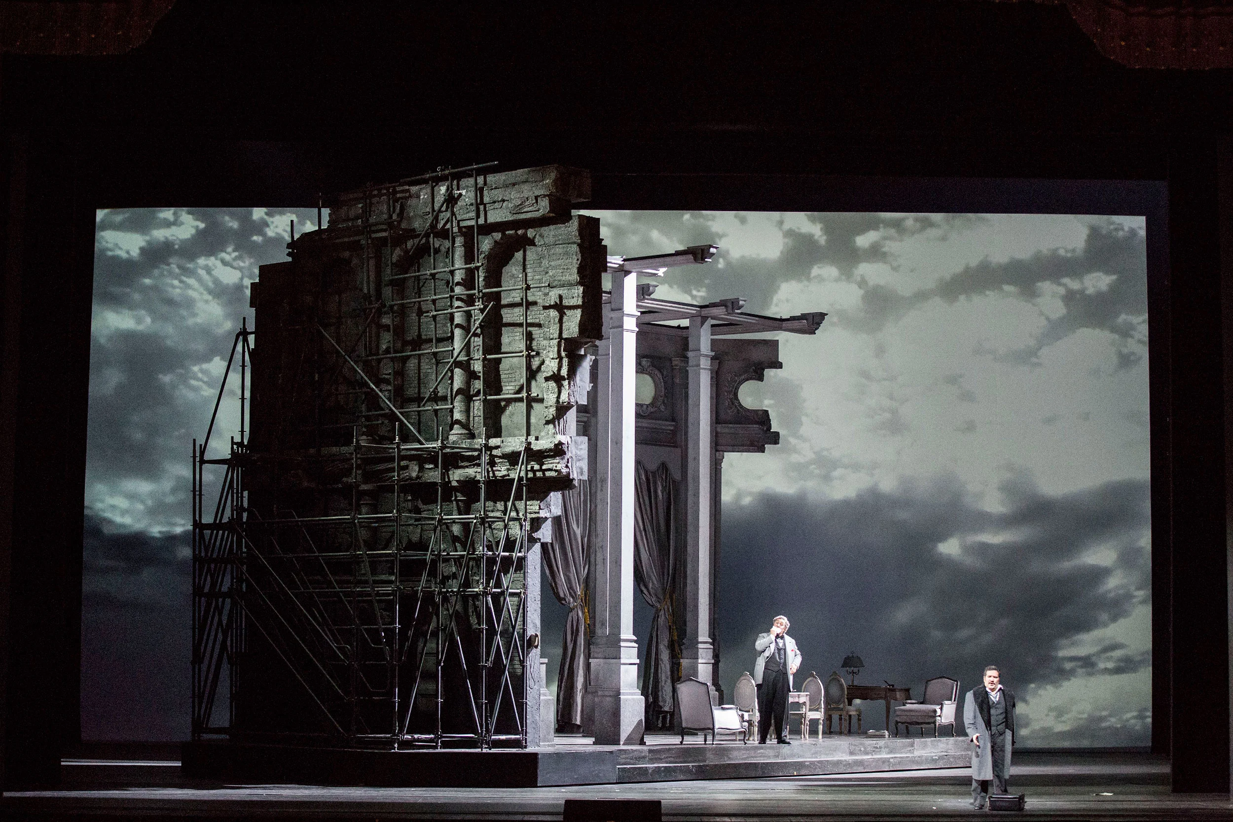 Scaffolded ruin and columns on a turning stage against a grey cloudy backdrop, two singers – Don Pasquale at La Scala, Milan, Italy, 2018 – opera set design