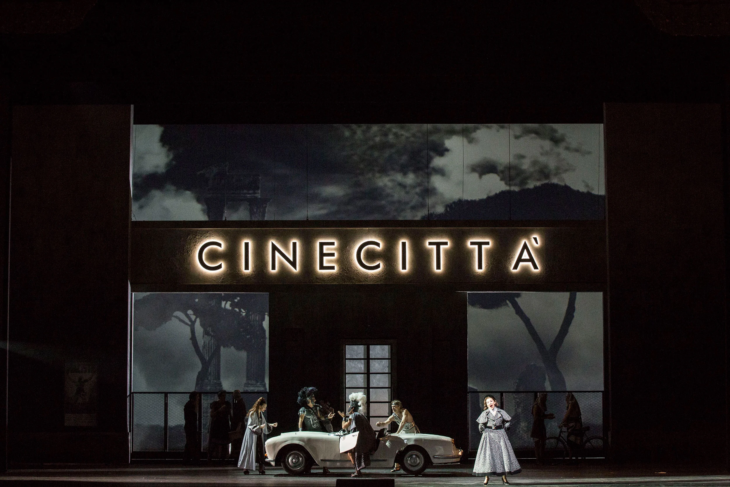 Illuminated Cinecitta sign above a building front, singers around a white convertible – Don Pasquale at La Scala, Milan, Italy, 2018 – opera set design