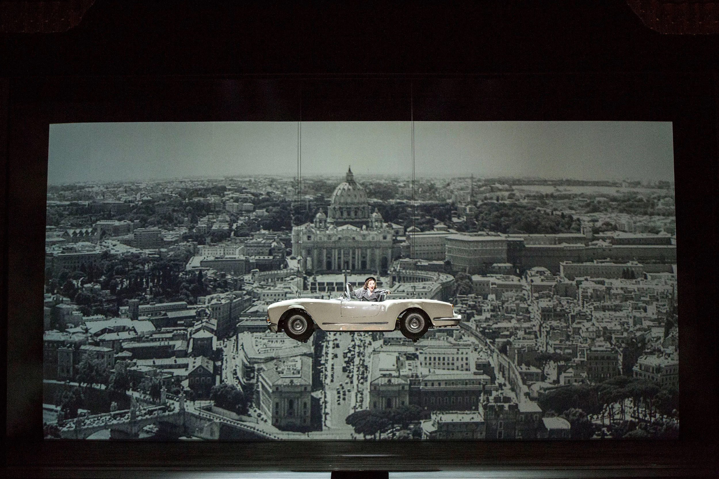 Singer driving a white convertible suspended above a black and white aerial view of Rome – Don Pasquale at La Scala, Milan, Italy, 2018 – opera set design