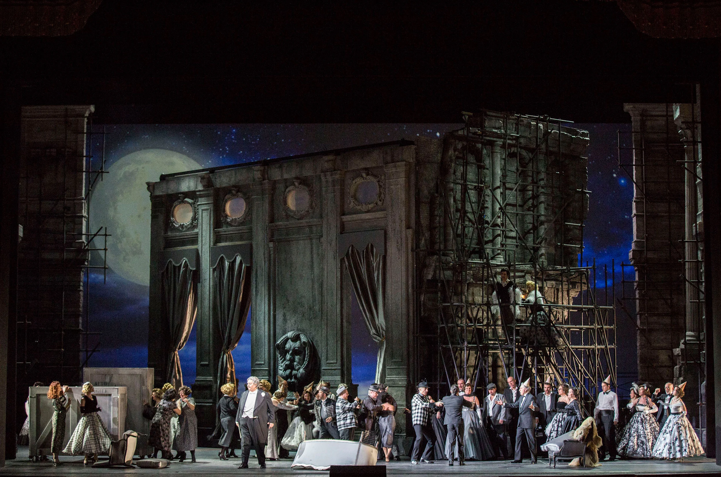 Full cast in front of a dark palace facade and scaffolding under a blue moonlit sky – Don Pasquale at La Scala, Milan, Italy, 2018 – opera set design