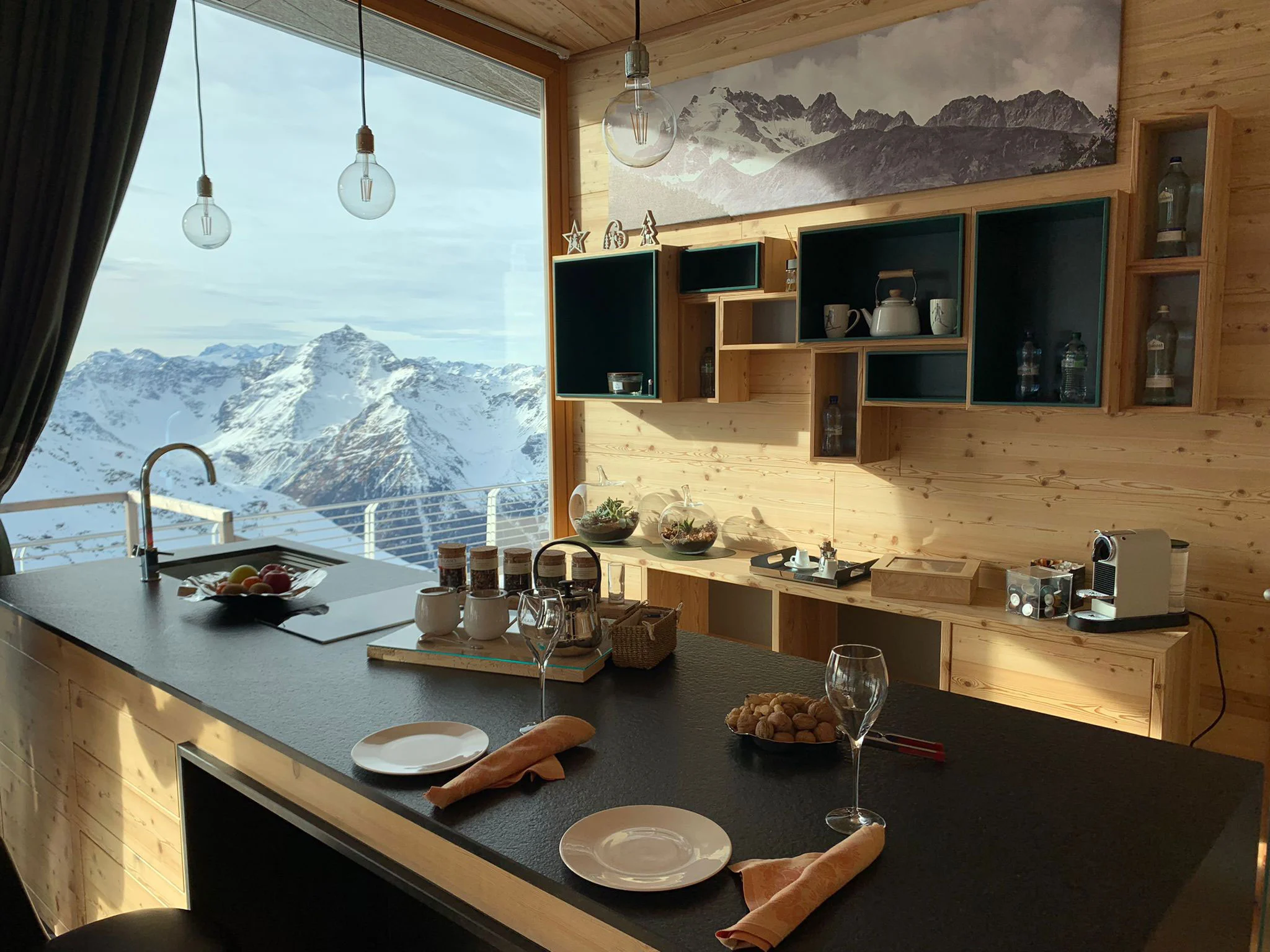 Kitchen bar in pale wood and dark stone, set for breakfast, with a snowy peak framed by the window – Levissima 3000 Chalet, Valdisotto, Italy, 2018 – architecture