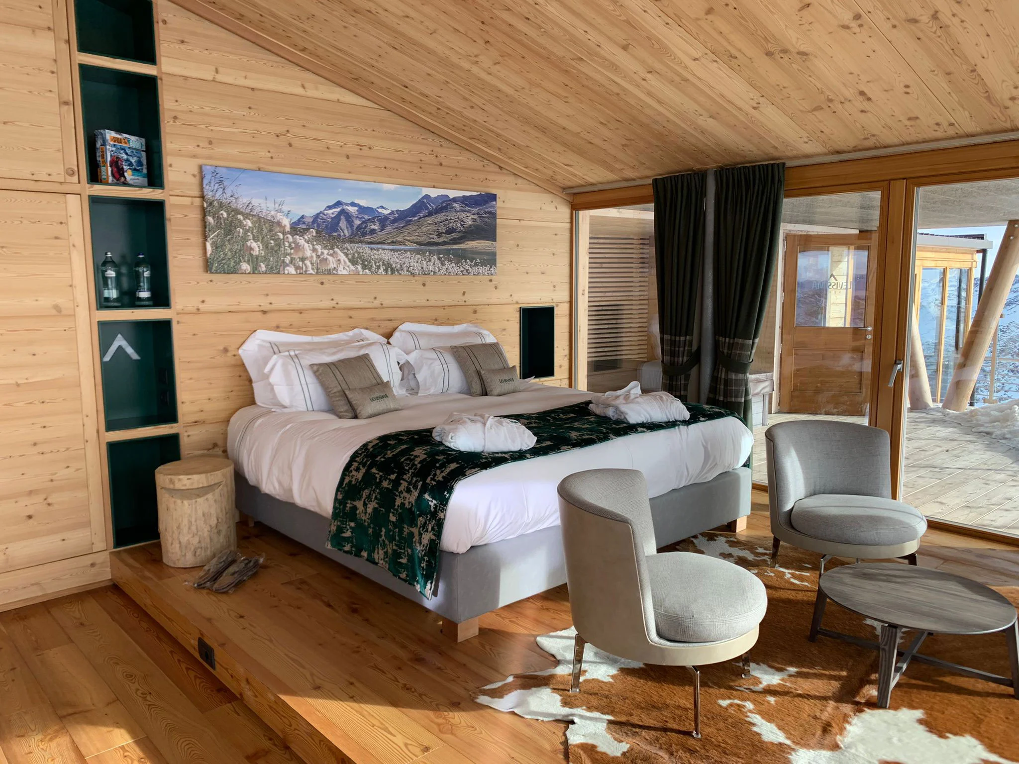 Wood-lined bedroom with a double bed on a raised platform, a mountain photograph above it and two armchairs – Levissima 3000 Chalet, Valdisotto, Italy, 2018 – architecture
