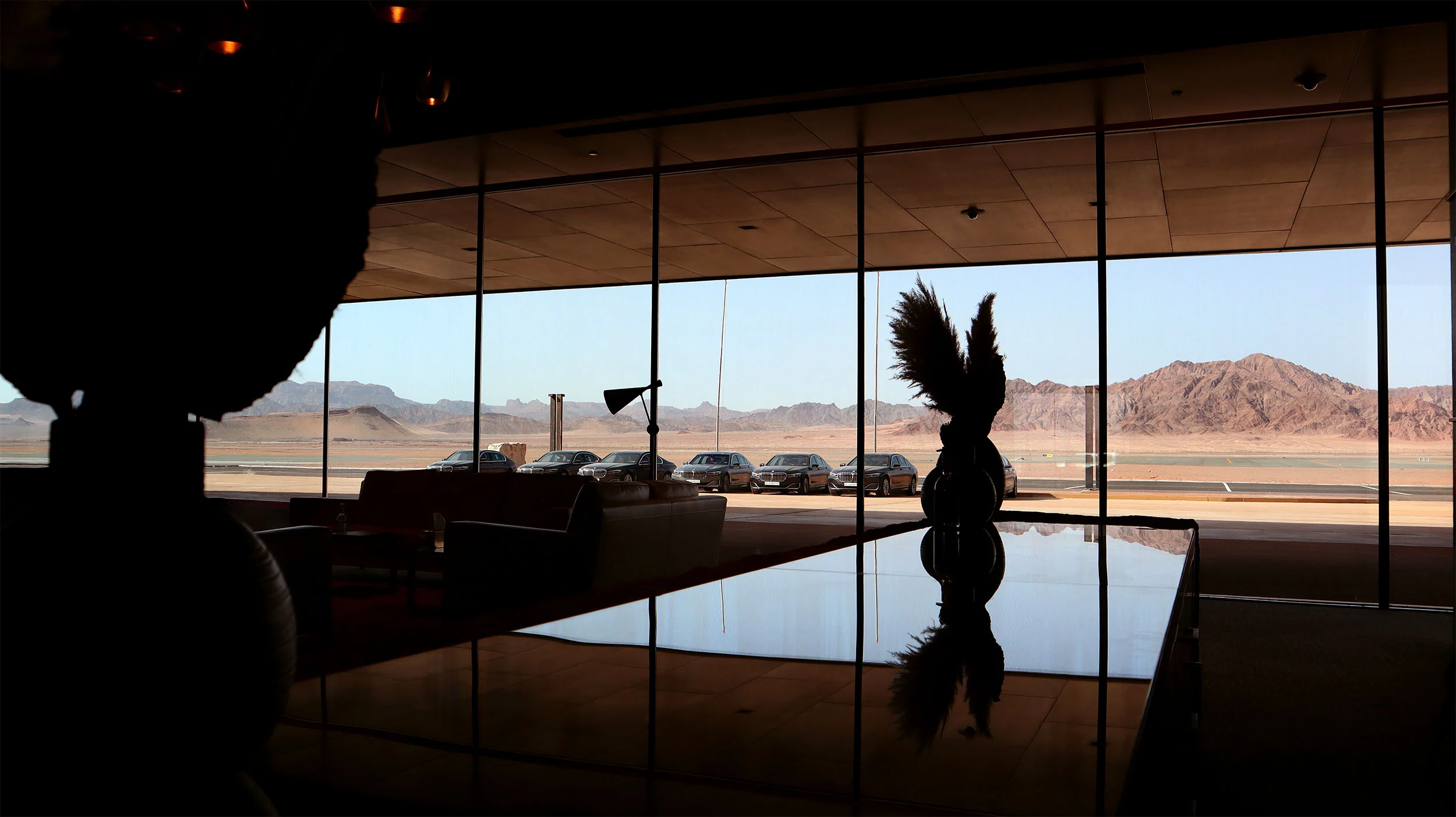 Lounge interior in silhouette, a glossy table reflecting a palm sculpture before mountain views – Prince Abdul Majeed bin Abdulaziz Airport, AlUla, Saudi Arabia, 2021 – architecture