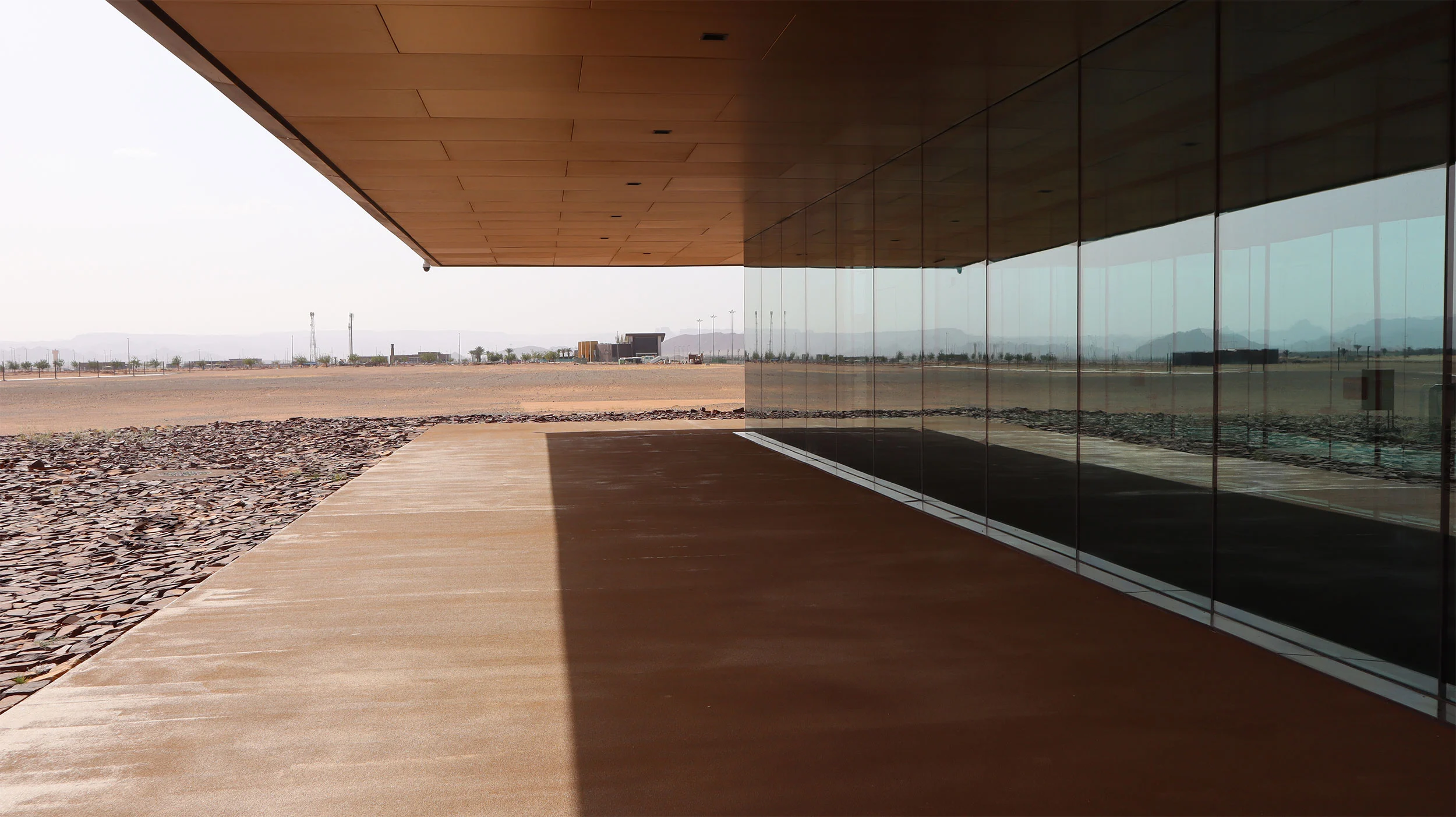 Shaded walkway beneath the cantilevered roof, the mirrored glass facade reflecting the desert – Prince Abdul Majeed bin Abdulaziz Airport, AlUla, Saudi Arabia, 2021 – architecture