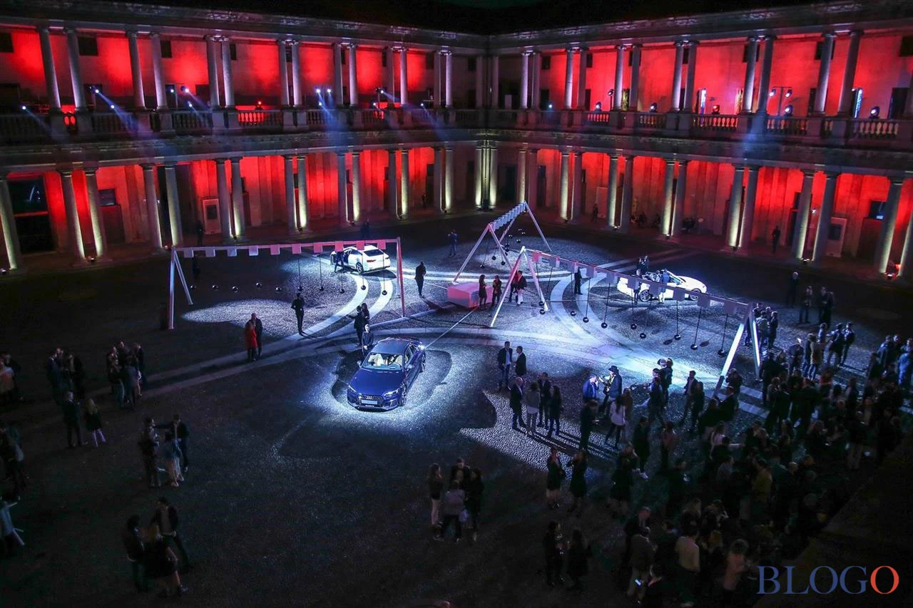 Palazzo courtyard in red light with Audi cars on lit platforms, swing frames and visitors at night – Audi City Lab at Fuorisalone, Milan, Italy, 2017 – event design