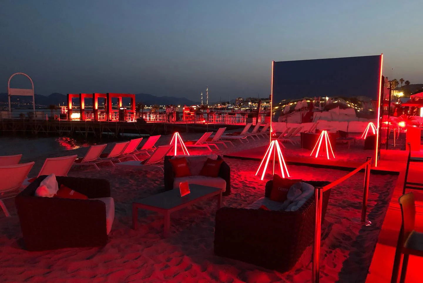 Beach lounge at dusk with sofas, red neon tripods and a large mirror panel standing on the sand – Campari at the 75th Cannes Film Festival, France, 2022 – event design
