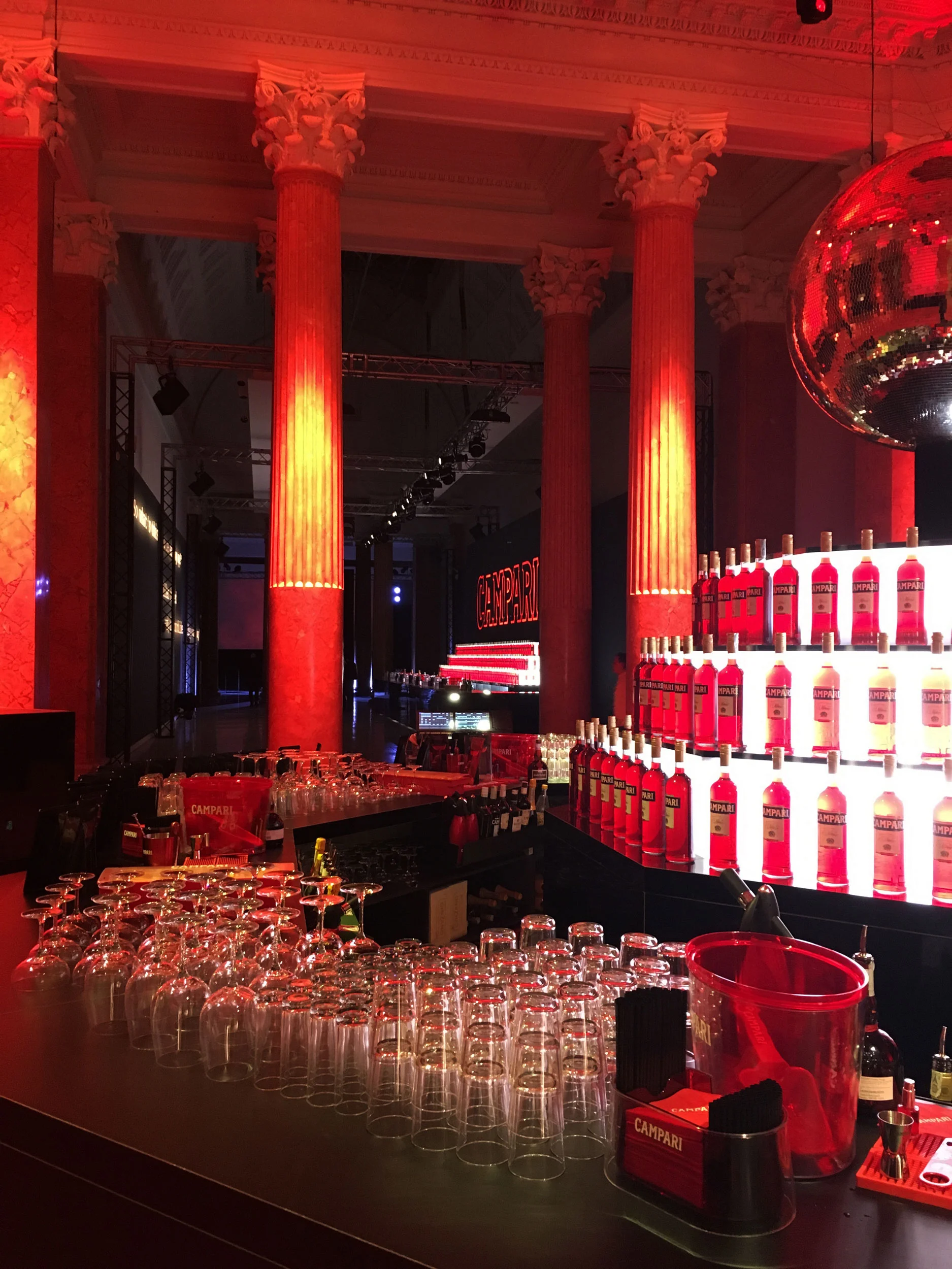 Bar counter with rows of glasses and backlit Campari bottles between red-lit classical columns – Campari Red Diaries, Milan, Italy, 2017 – event design