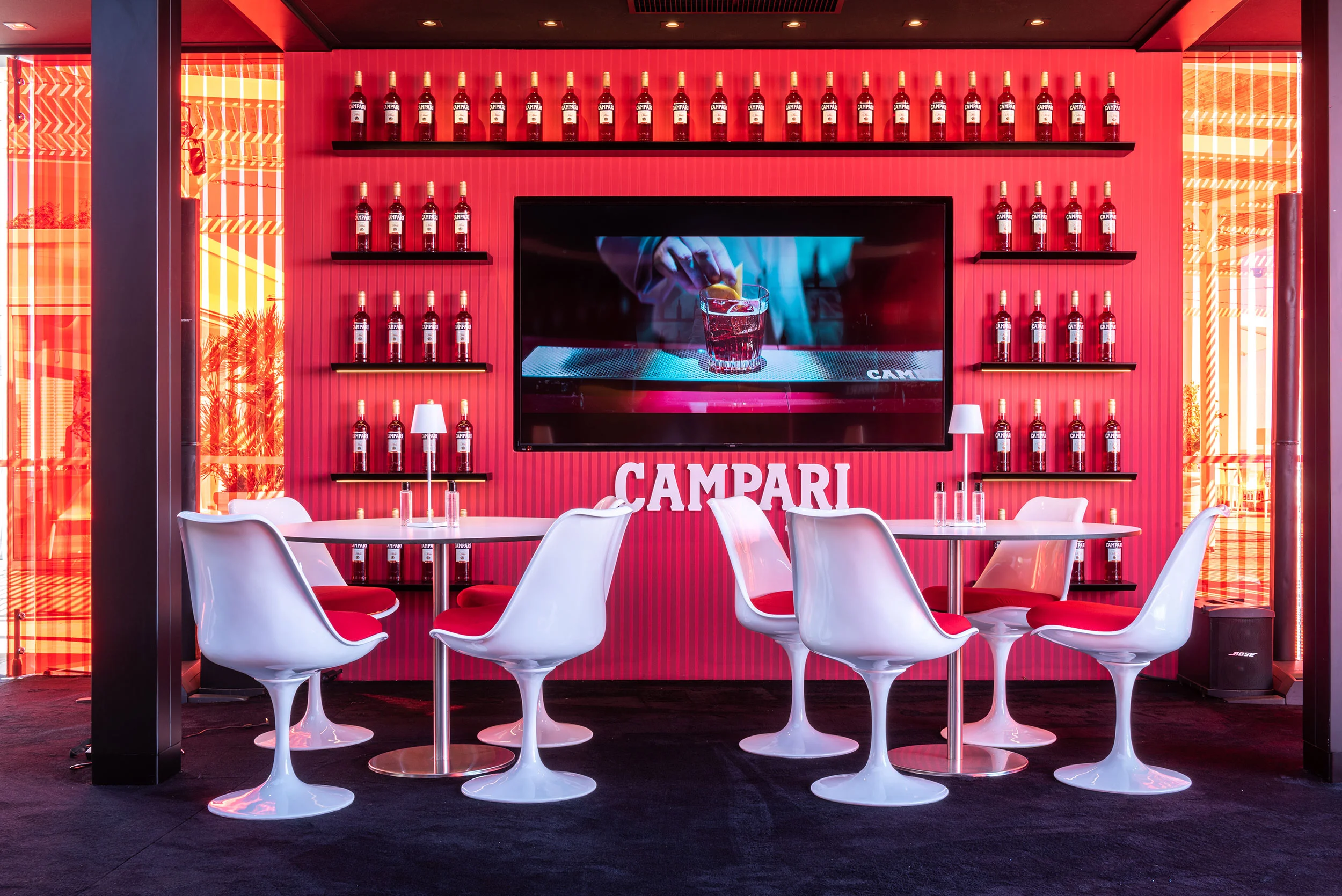 Lounge corner with white tulip chairs before a red wall of Campari bottles and a video screen – Campari Lounge Experience at Cannes Film Festival, France, 2022 – event design