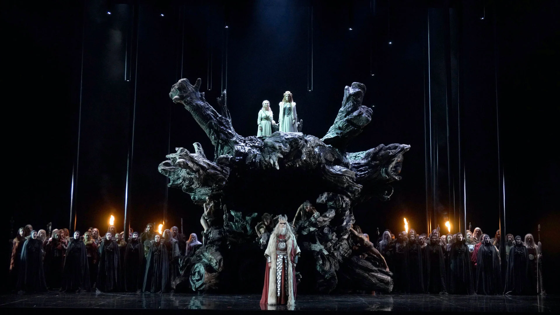 Huge blackened tree root at centre stage with two women in pale gowns on top and the chorus lined up below – Norma at Teatro Real, Madrid, Spain, 2016 – opera set design