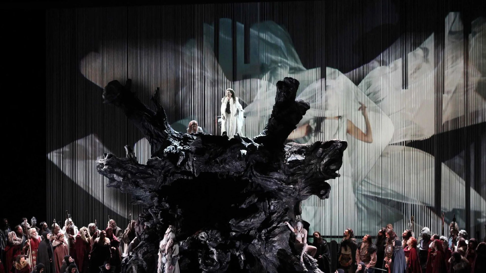Dark sculpted tree root set with a figure in white on top, against a projection of billowing white cloth – Norma at Teatro Real, Madrid, Spain, 2016 – opera set design