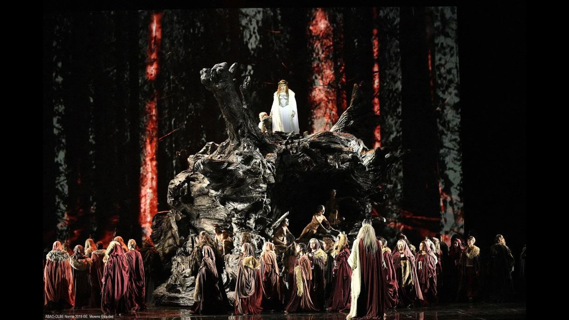 Norma staged at Teatro Real: a priestess in white atop a giant gnarled tree trunk, chorus below in red robes – Norma at Teatro Real, Madrid, Spain, 2016 – opera set design
