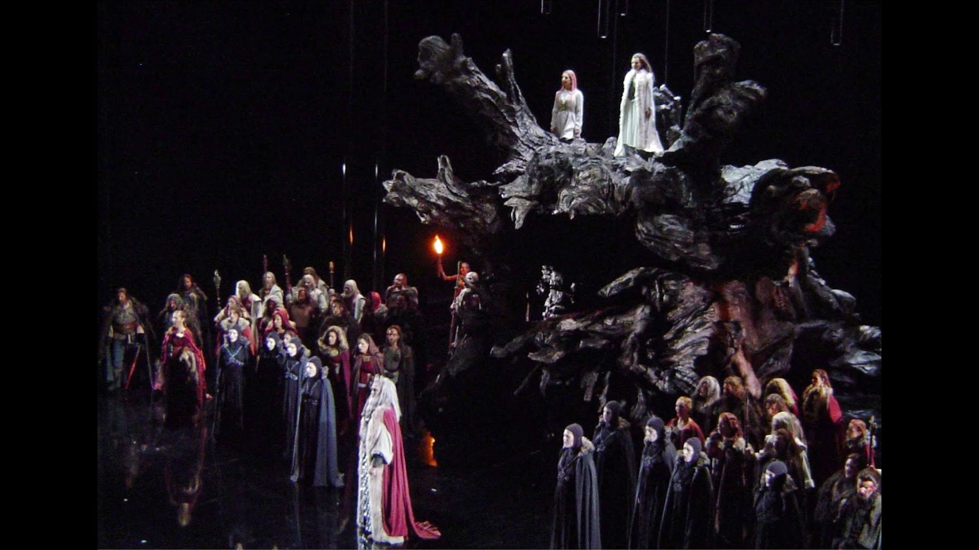 Side view of the tree-root set with two figures on top and the hooded chorus holding torches – Norma at Teatro Real, Madrid, Spain, 2016 – opera set design