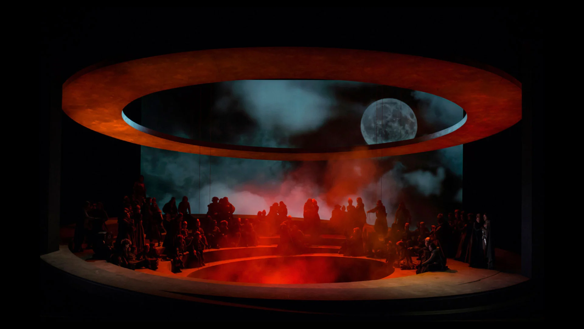 Circular tiered stage in red light and smoke beneath an orange ring, with a projected moon and clouds – Otello at Teatro Carlo Felice, Genoa, Italy, 2014 – opera set design