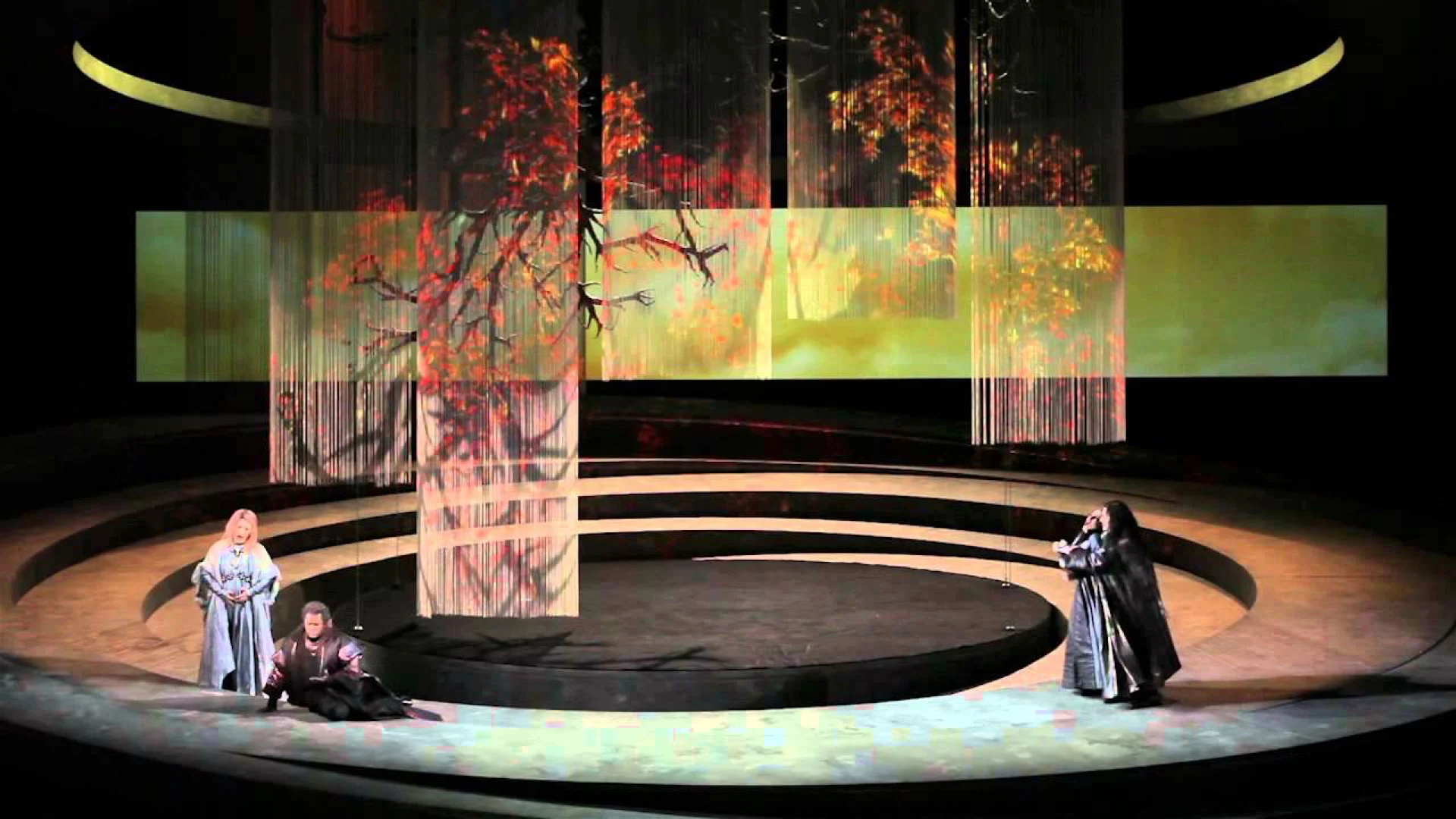 Three singers on the circular stage beneath string curtains showing an autumn tree in red and gold – Otello at Teatro Carlo Felice, Genoa, Italy, 2014 – opera set design