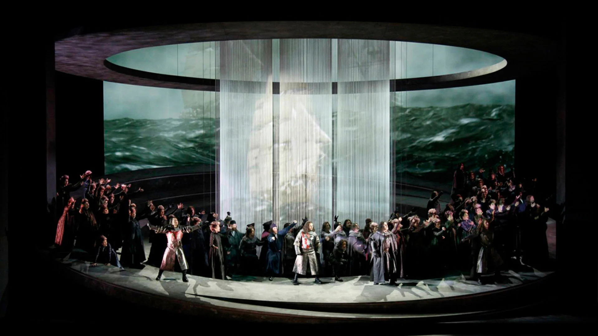 Chorus on the circular stage before string curtains projecting a sailing ship on a stormy sea – Otello at Teatro Carlo Felice, Genoa, Italy, 2014 – opera set design