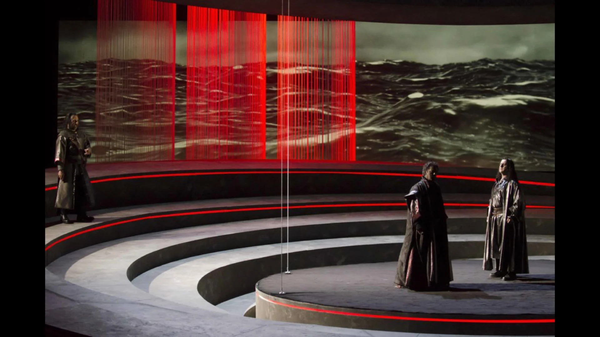 Three performers on the curved tiers before a stormy sea projection crossed by red string curtains – Otello at Teatro Carlo Felice, Genoa, Italy, 2014 – opera set design