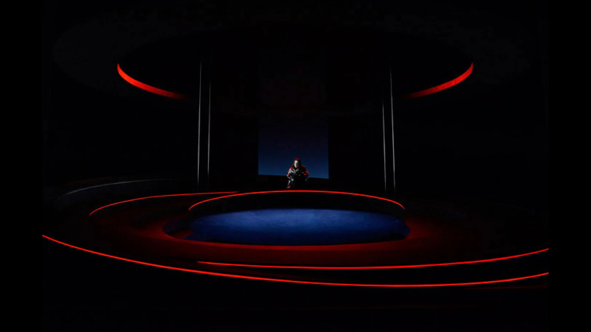 Dark stage with a lone figure seated at the centre, outlined by red light rings on the tiers – Otello at Teatro Carlo Felice, Genoa, Italy, 2014 – opera set design