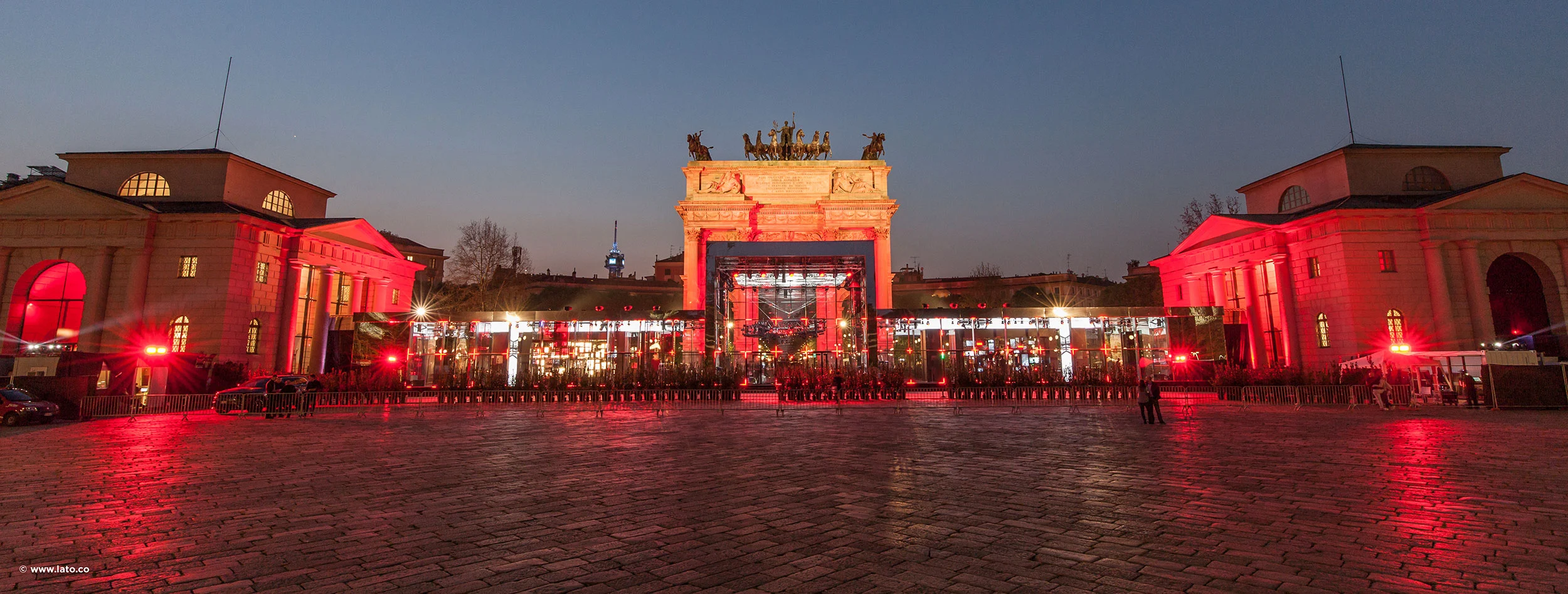 Wide dusk view of the square and the arch, with long red-lit pavilions and flanking buildings – Cartier The Legendary Thrill, Milan, Italy, 2018 – event design