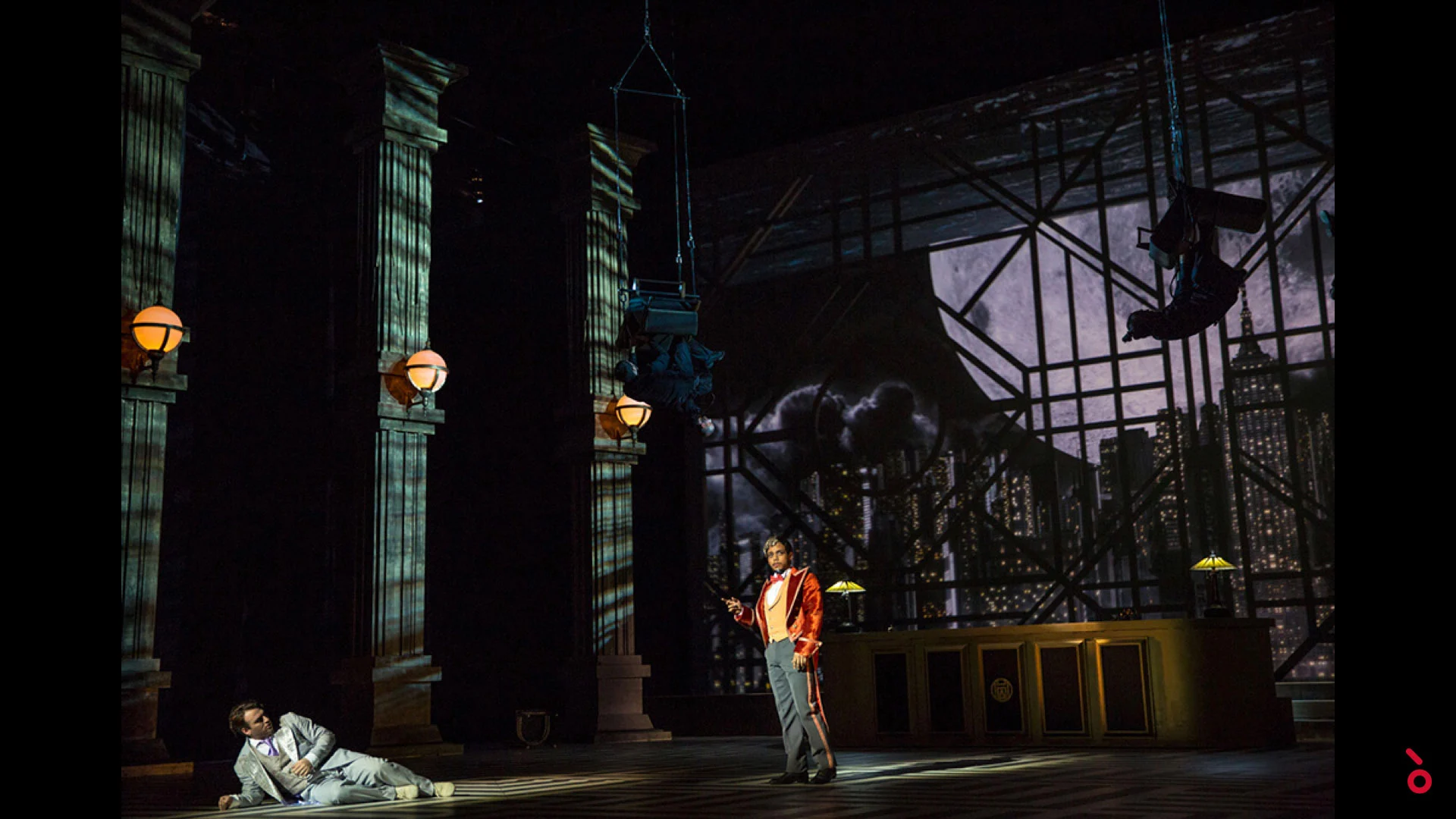 Two performers in a dim lobby set of tall columns and globe lamps, a night city skyline behind – The Opera! at the Royal Opera House Muscat, Oman, 2019 – set design