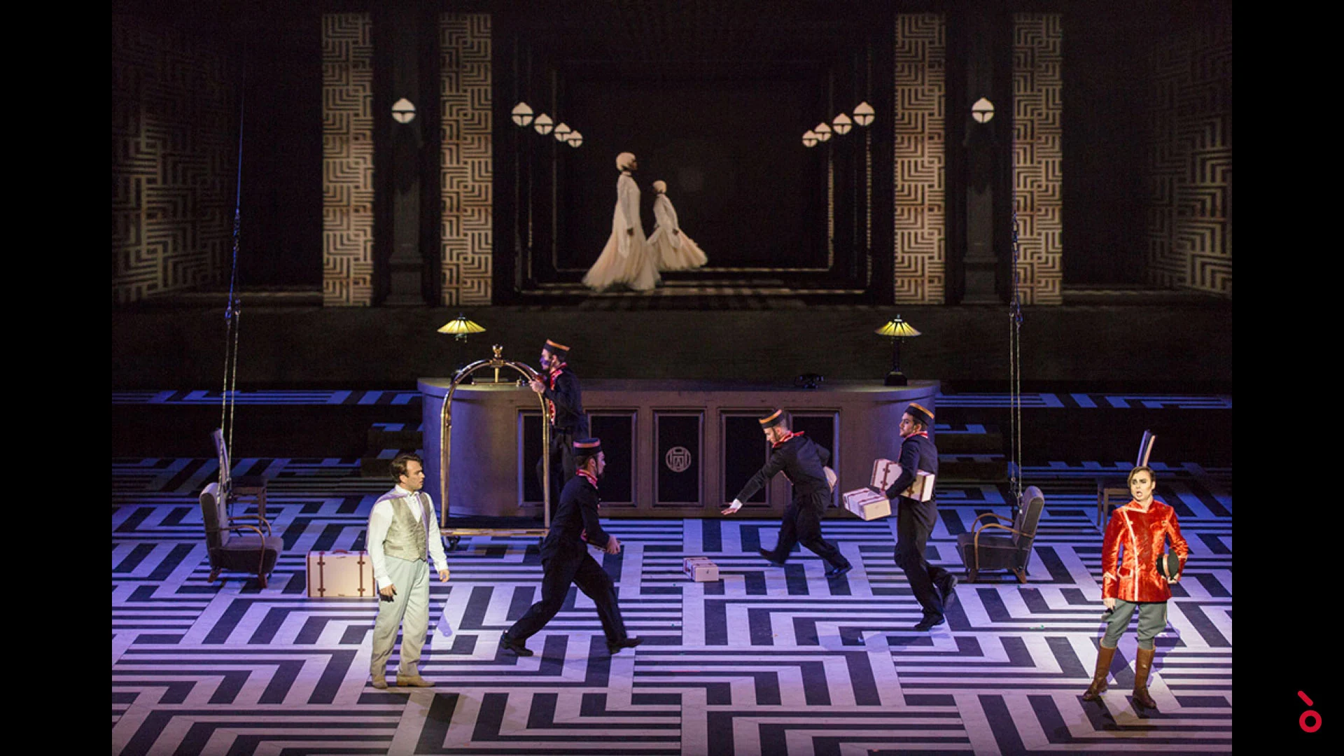 Bellhops carrying luggage across the hotel set while two women in white walk along a rear gallery – The Opera! at the Royal Opera House Muscat, Oman, 2019 – set design