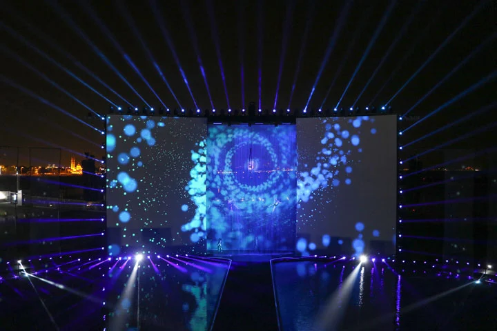 Stage at night framed by blue light rays, with projected bubbles and a vortex image across the screens – Dubai Water Canal Opening Ceremony, United Arab Emirates, 2016 – event design