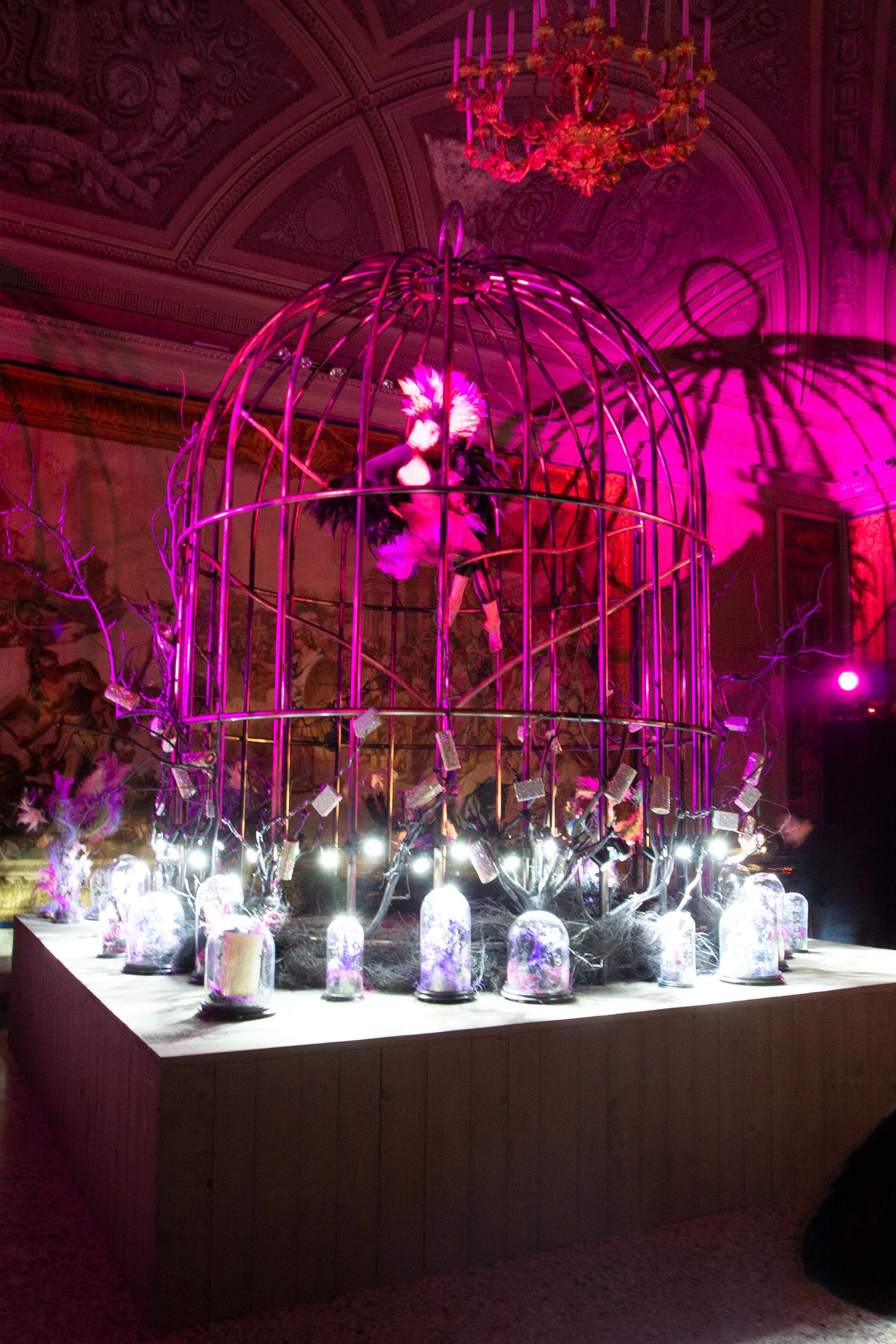 Giant birdcage on a wooden plinth under magenta light, with a feathered figure inside and jewels displayed in glass domes – Swarovski Fashionation Party at Palazzo Reale, Milan, Italy, 2011 – event design