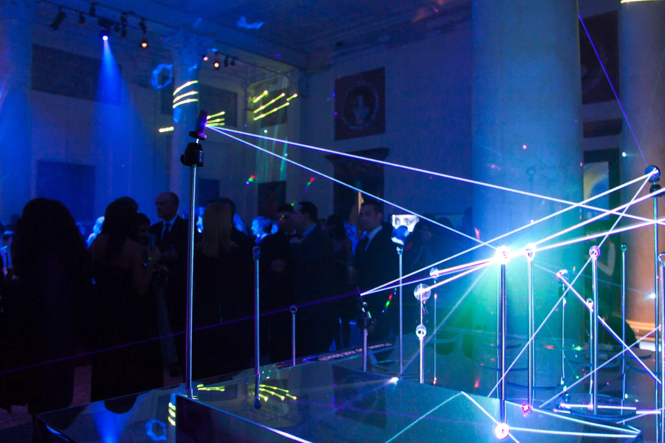 Guests in a blue-lit hall crossed by laser lines strung between thin metal poles on a mirrored base – Swarovski Fashionation Party at Palazzo Reale, Milan, Italy, 2011 – event design