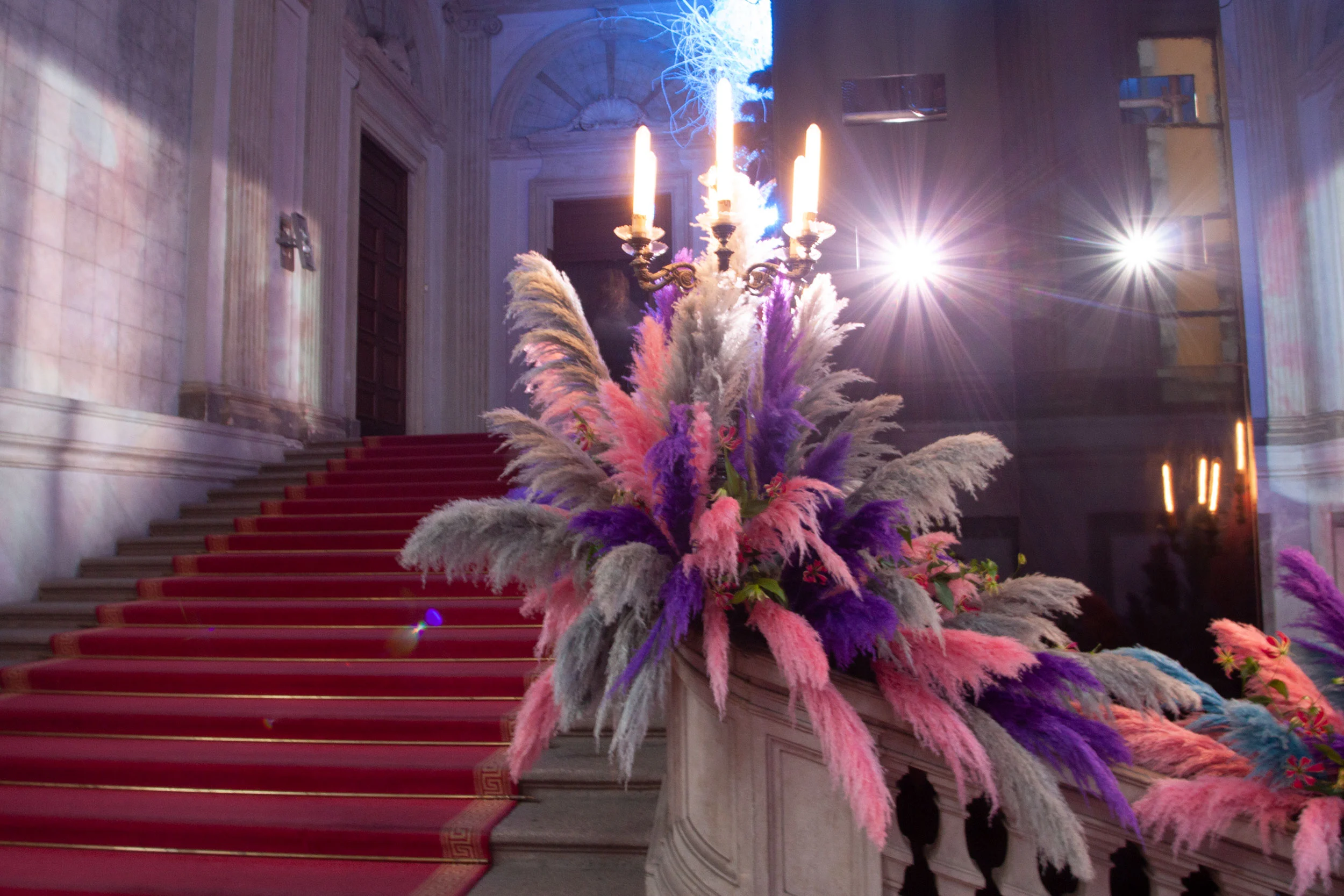 Red-carpeted grand staircase with candelabra and plumes of pink, purple and grey feathers on the balustrade – Swarovski Fashionation Party at Palazzo Reale, Milan, Italy, 2011 – event design