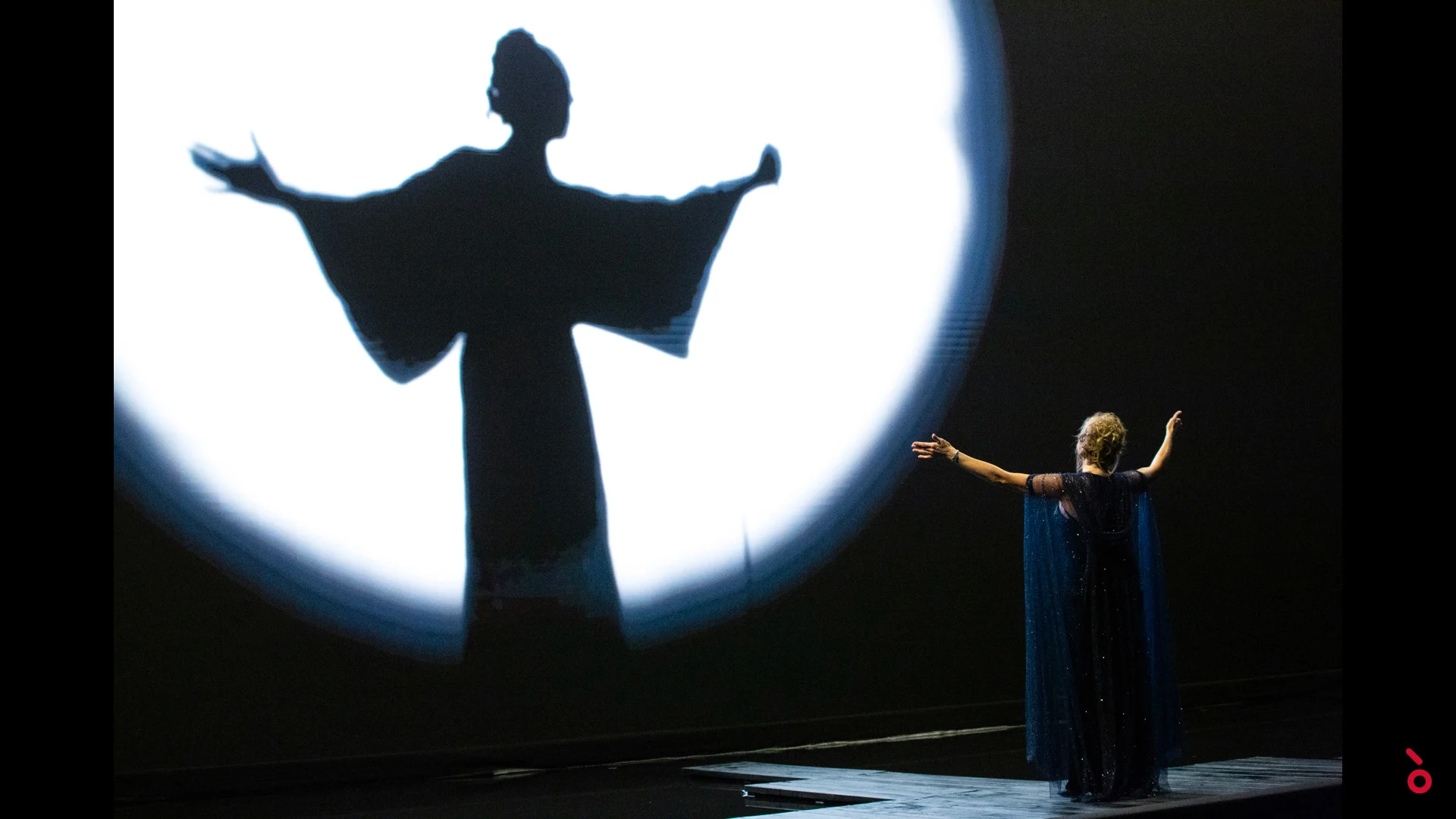 Singer in a blue cape with open arms facing her giant shadow in a circle of white light – A Riveder le Stelle at La Scala, Milan, Italy, 2020 – opera set design