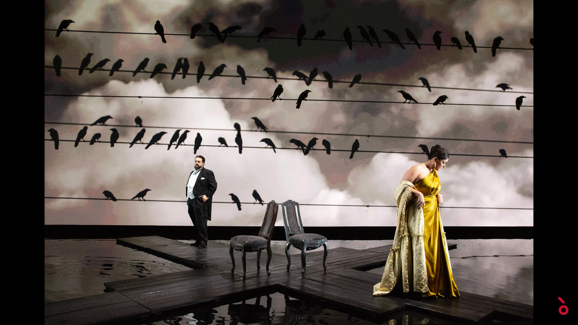 Two singers by empty chairs on a wet wooden deck, crows on wires projected across a cloudy sky – A Riveder le Stelle at La Scala, Milan, Italy, 2020 – opera set design