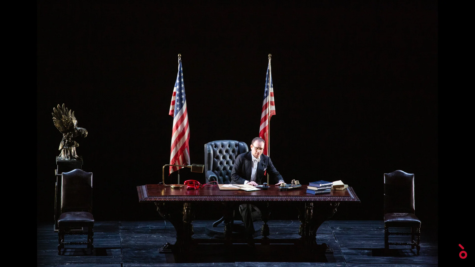 Actor seated at a large desk between two American flags, an eagle sculpture at left – A Riveder le Stelle at La Scala, Milan, Italy, 2020 – opera set design