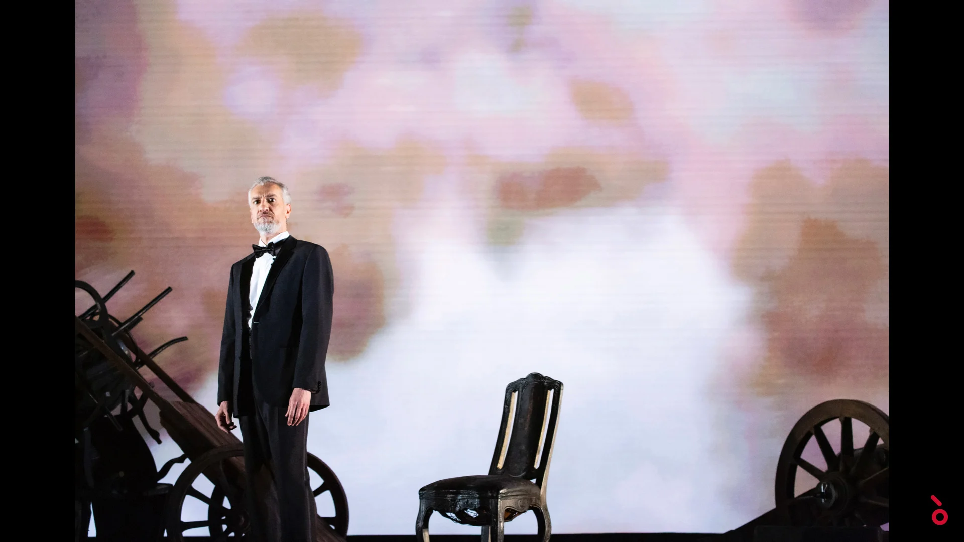 Singer in black tie beside an empty chair and old cart wheels, soft pink clouds projected behind – A Riveder le Stelle at La Scala, Milan, Italy, 2020 – opera set design