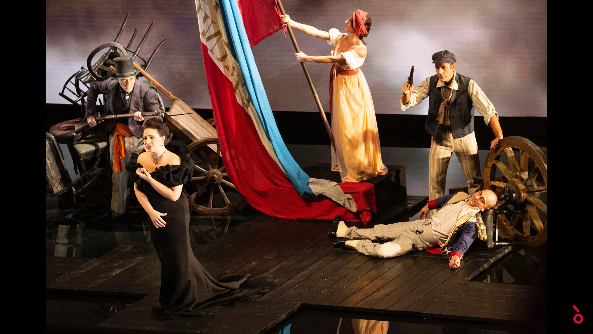 Singer in a black gown before a barricade scene with a large tricolour flag, cart wheels and actors – A Riveder le Stelle at La Scala, Milan, Italy, 2020 – opera set design