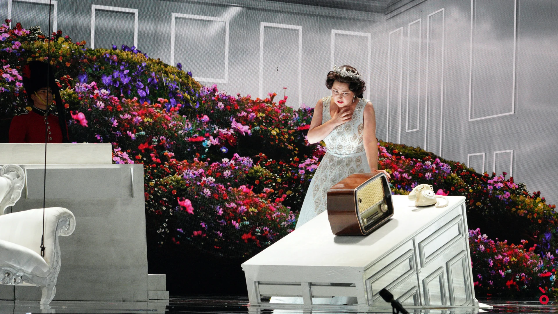 The queen in a white gown beside an old radio on a white chest, against a wall of colourful flowers – Elisabetta, Regina d'Inghilterra at Rossini Opera Festival, Pesaro, Italy, 2021 – set design