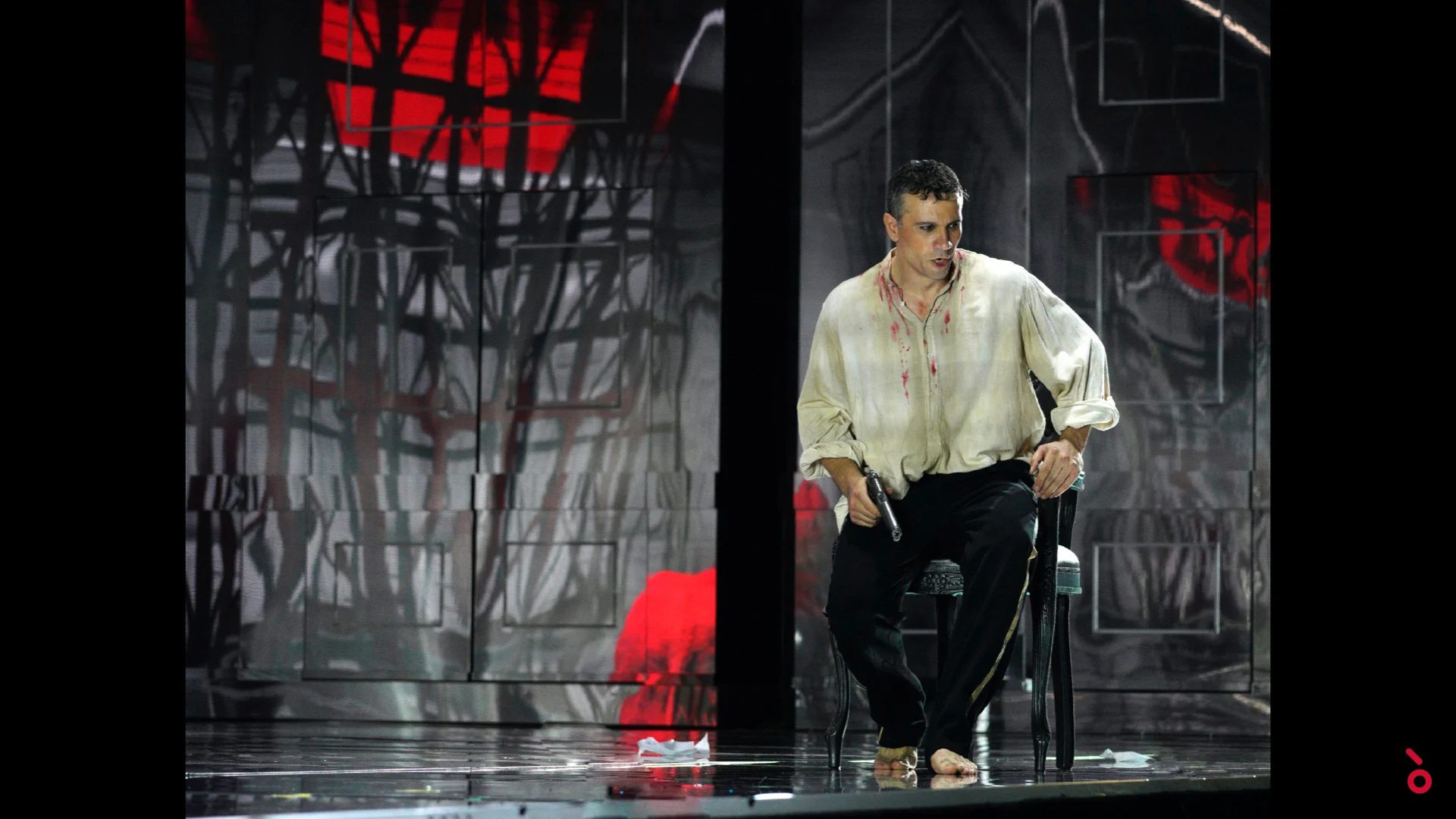 Singer in a bloodstained shirt seated on a chair, bare trees and red light projected behind – Elisabetta, Regina d'Inghilterra at Rossini Opera Festival, Pesaro, Italy, 2021 – set design