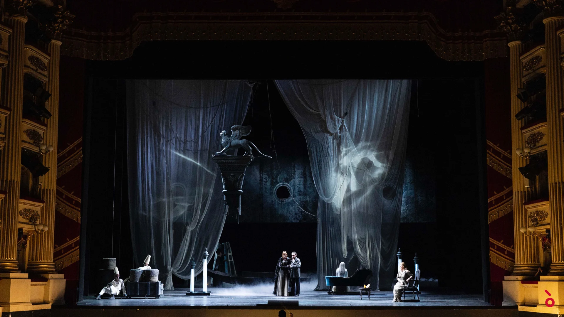 Intimate scene framed by sheer grey curtains with projections, a winged lion statue hanging above the stage – La Gioconda at La Scala, Milan, Italy, 2022 – opera set design