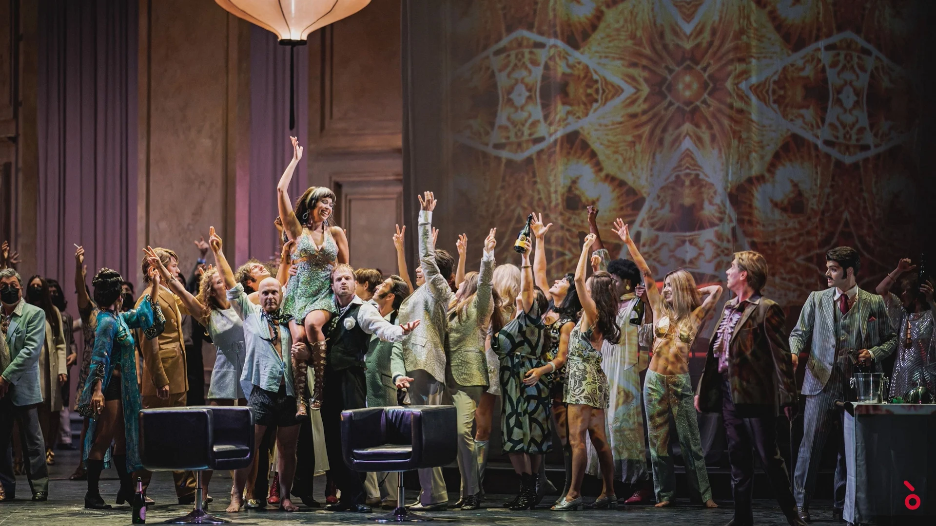 Party scene with the chorus in 1960s costumes raising their arms under a paper lantern, kaleidoscopic projection behind – La Traviata at Maggio Musicale Fiorentino, Florence, Italy, 2022 – opera set design